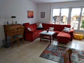 Un salon avec un canapé rouge, un bureau en bois et une petite table. Une fenêtre offre une vue sur les bâtiments à l’extérieur.