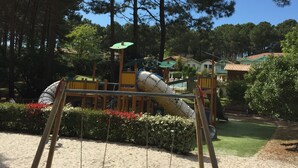 A playground with swings and a slide, surrounded by trees and a sandy area. - House 6/8 pers in holiday resort - pine forest, ocean, lake, your choice! (Lacanau Océan)