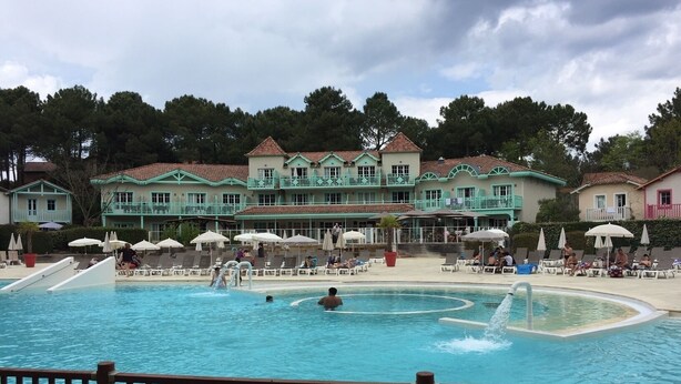 A swimming pool area with lounge chairs and umbrellas, surrounded by buildings with a consistent architectural style. - House 6/8 pers in holiday resort - pine forest, ocean, lake, your choice! (Lacanau Océan)