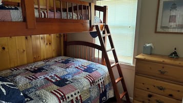 Uma cama beliche com colcha, cómoda de madeira e janela com persianas.
