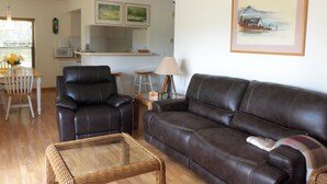 Una sala de estar con un sofá de cuero marrón, una mesa de centro de mimbre, un sillón de cuero negro y un comedor con una silla y una mesa blancos.