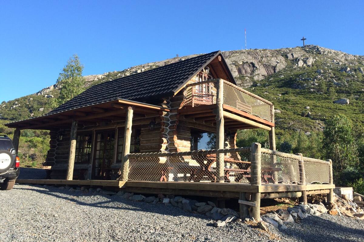A rustic log cabin with a balcony, surrounded by a gravel driveway and a mountainous backdrop.