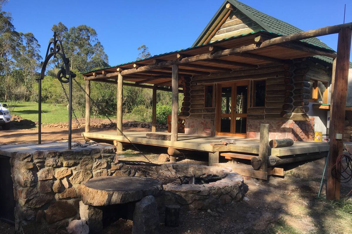 A rustic cabin with a wooden porch, stone walls, and a stone fire pit.