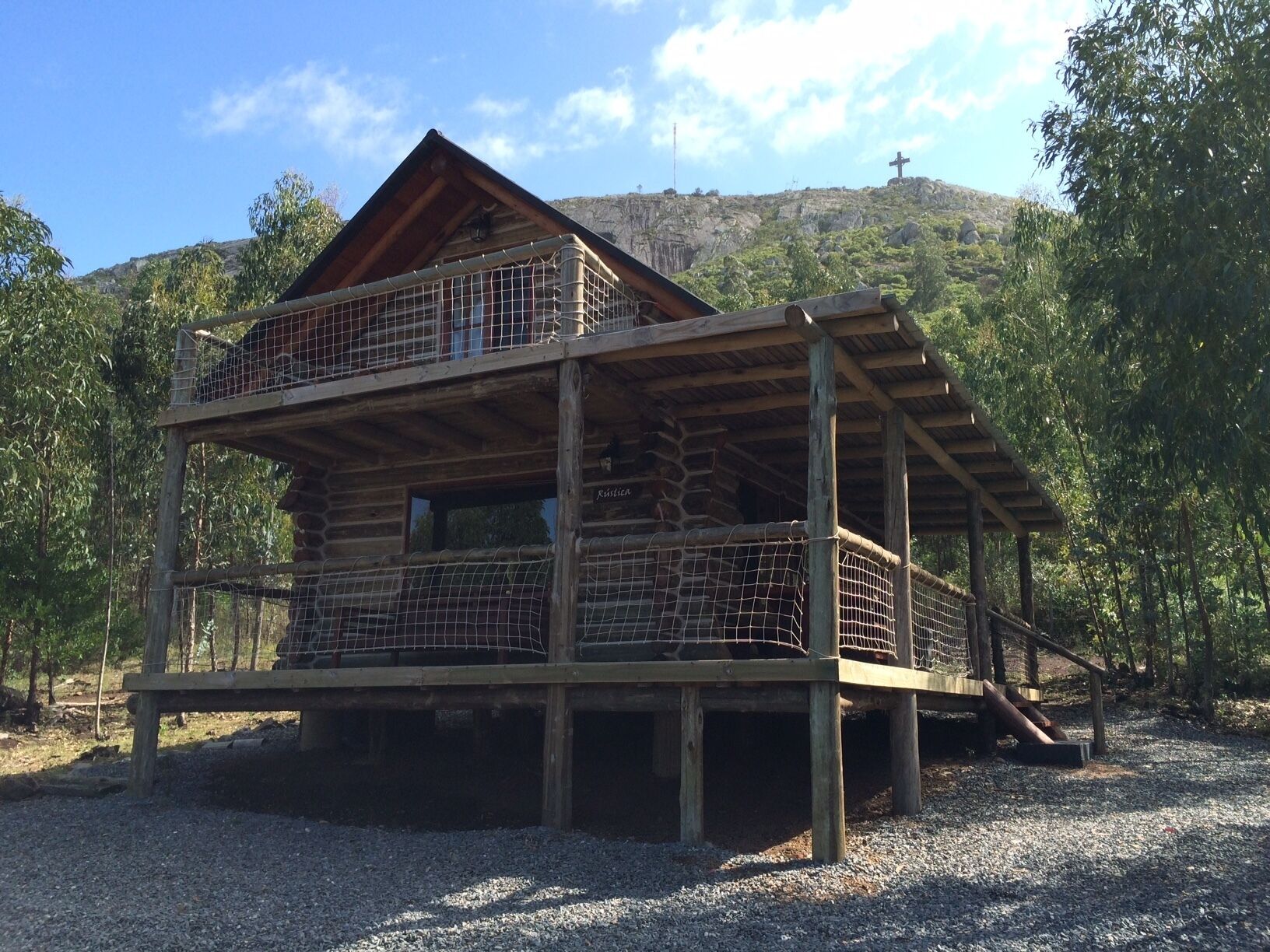 Una baita in legno a due piani con un balcone, circondata da alberi e con una collina sullo sfondo.