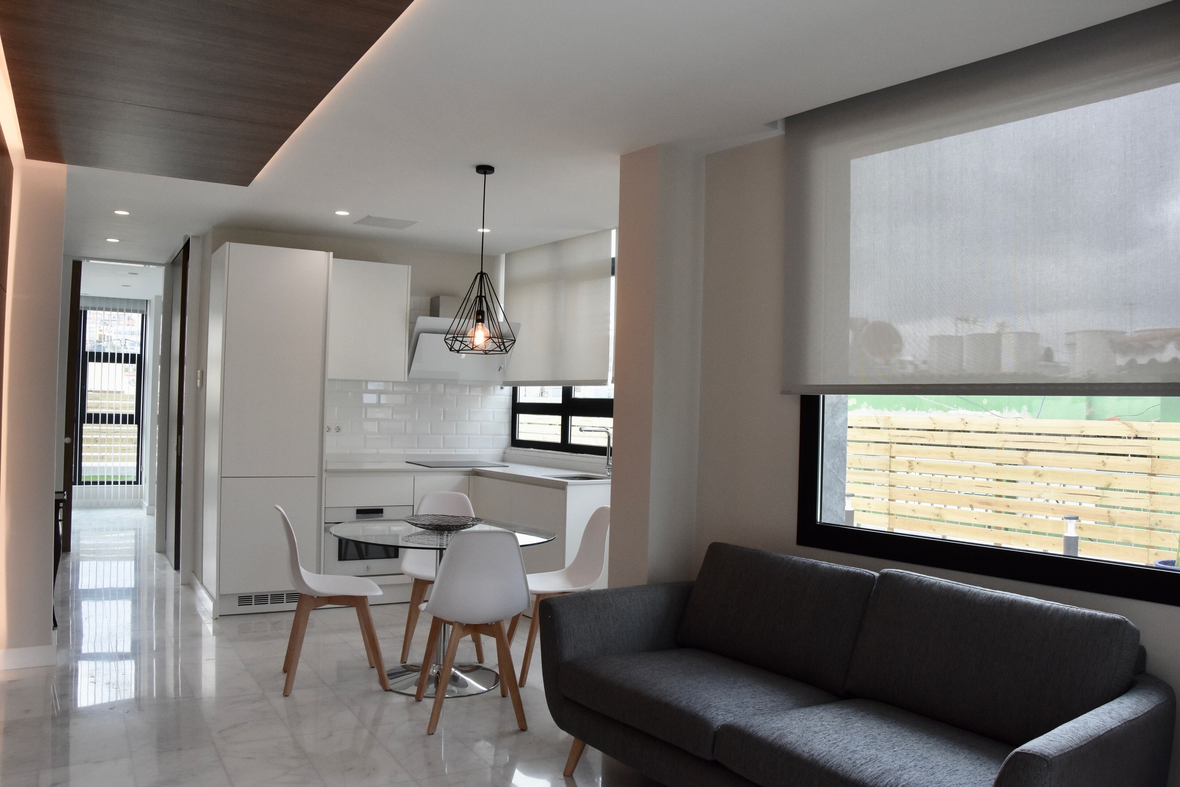 Una cocina moderna con armarios blancos, un comedor con mesa y sillas, y un sofá gris cerca de un ventanal grande.