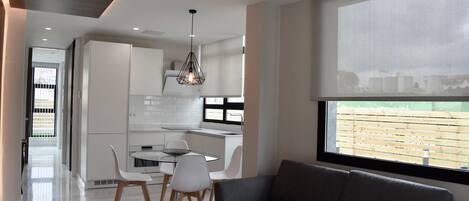 Una cocina moderna con armarios blancos, un comedor con mesa y sillas, y un sofá gris cerca de un ventanal grande.