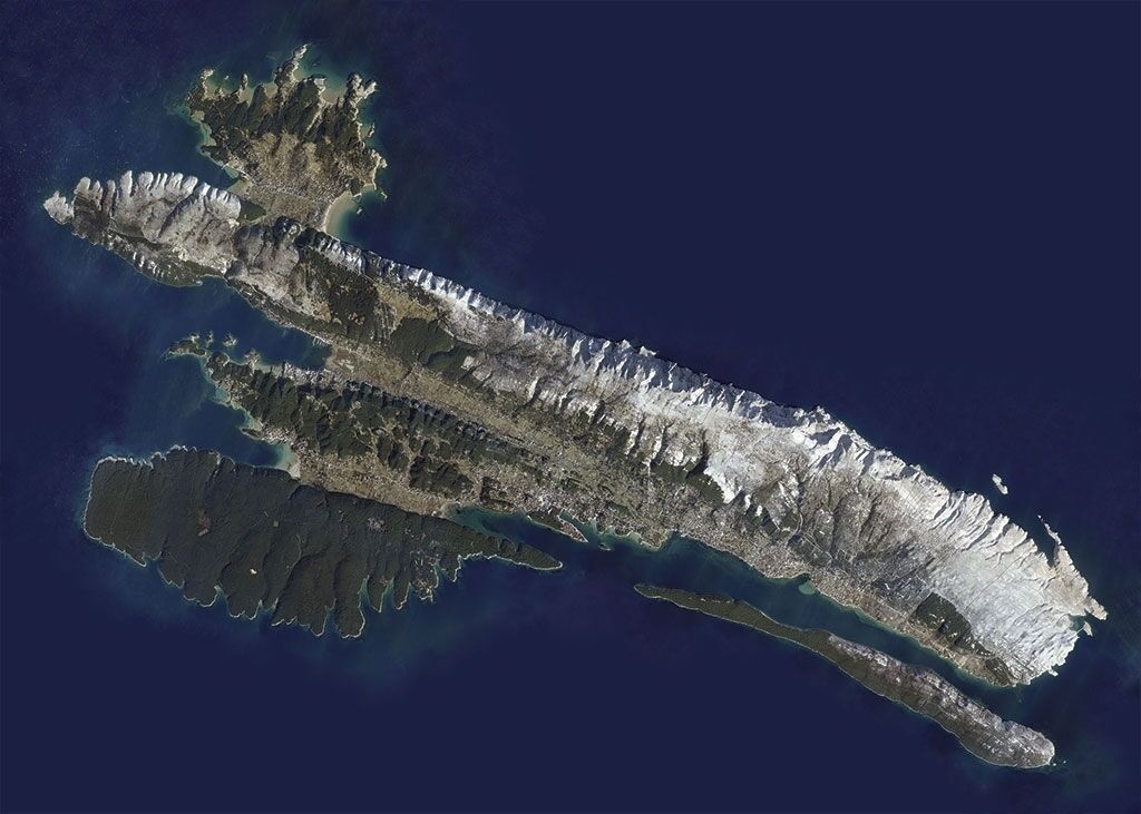 A satellite view of a long, narrow island with rugged terrain and a mix of mountainous and flat areas.