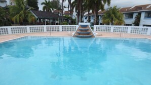 Ein Schwimmbecken mit mehrstufigem Wasserspiel, umgeben von einem weißen Zaun und Palmen.