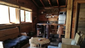 Interior rústico de una cabaña con un banco de madera, una mesa redonda de madera, un sofá, una kitchenette y una ventana con cortinas.