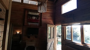 Interior de una cabaña de madera con chimenea, una lámpara en el techo y una ventana con vista a una terraza.
