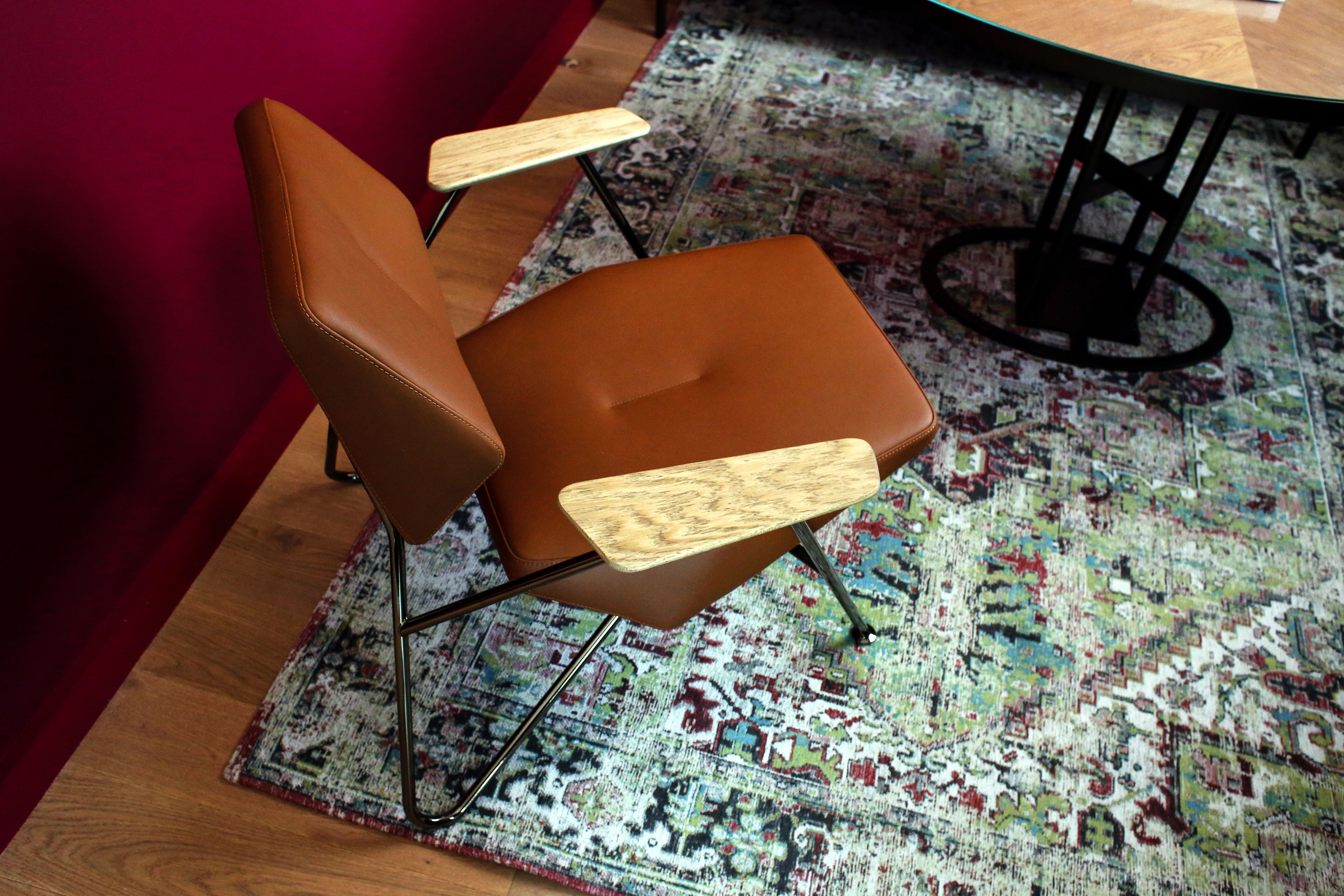 Una silla marrón con reposabrazos de madera y patas de metal, sobre una alfombra estampada.