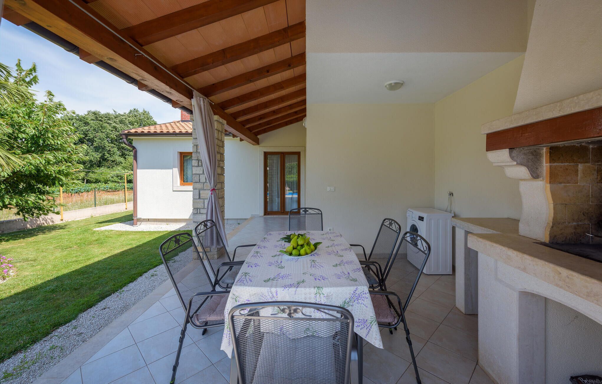 An outdoor patio with a table set for four, surrounded by chairs, a small kitchen area with a stone countertop, and a view of a garden and a building in the background.