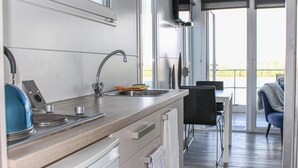 A modern kitchen with a stainless steel sink, a kettle, and a stove. There is a dining area with a table and chairs visible through a glass door.