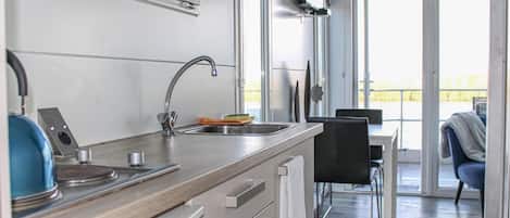 Une cuisine moderne équipée d’un évier en acier inoxydable, d’une bouilloire et d’une cuisinière. On aperçoit un coin repas avec une table et des chaises à travers une porte vitrée.