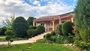 Ein Haus mit einem Ziegeldach, einer Veranda und einem gepflegten Garten mit verschiedenen Sträuchern und einer Rasenfläche.