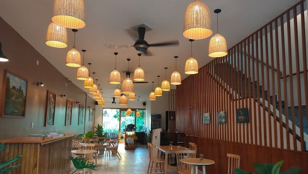 Interior de restaurant amb diverses làmpades de vimet penjants, taules i cadires de fusta, i una escala amb baranes de fusta.