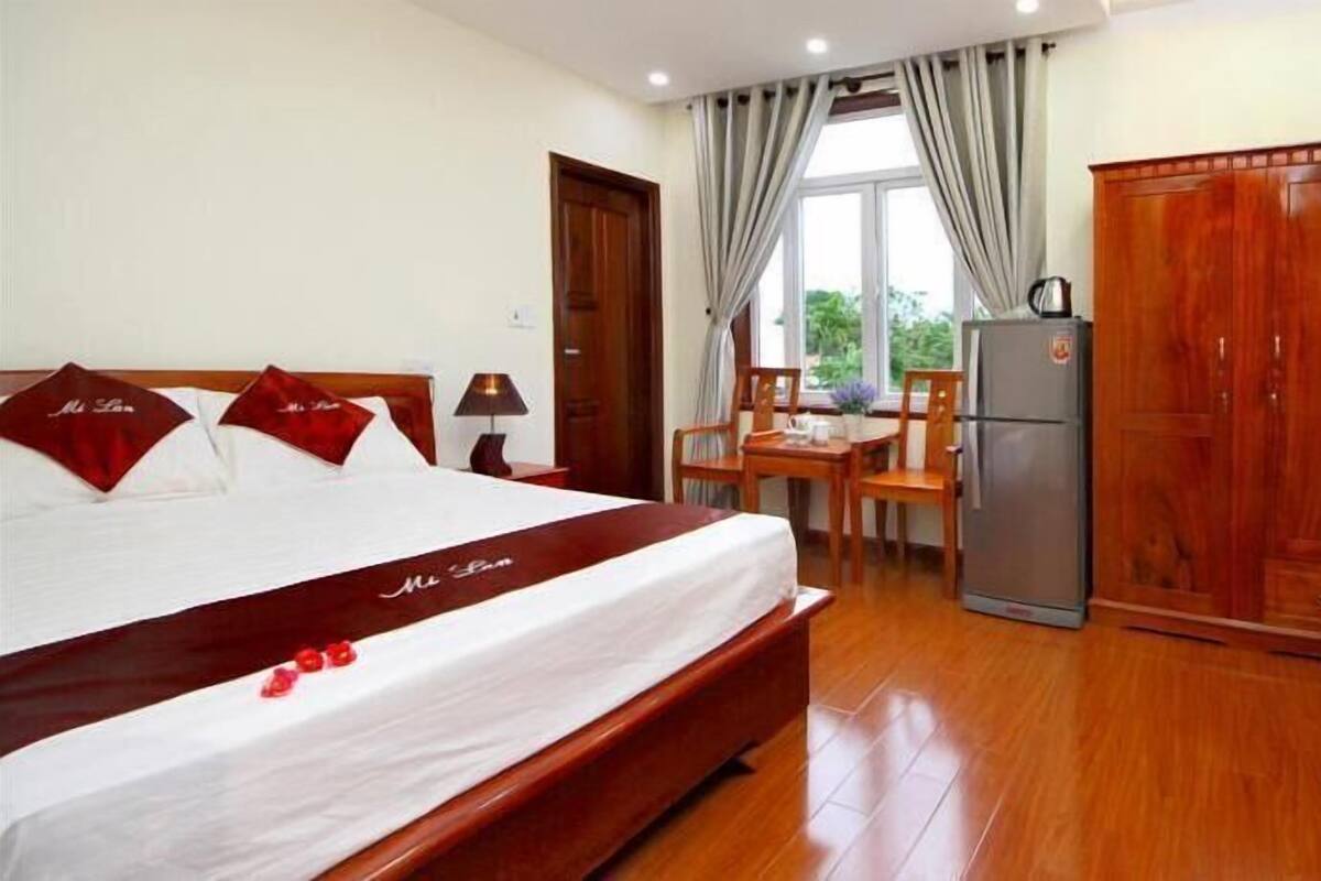 A hotel room with a large bed, a desk with a refrigerator, and a wardrobe.