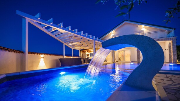 A swimming pool with a waterfall feature, a covered seating area, and a building with a lighted entrance. - Beach front home in Vrsi (Vrsi)
