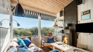 A cozy living room with a sofa, a table, and a fireplace. There is a TV mounted on the wall and a window with a view of the outdoors. - Nice home in Hvide Sande with WiFi (Hvide Sande)