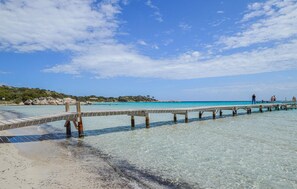 Ein Steg, der sich in klares türkisblaues Wasser erstreckt, mit einem Sandstrand und bewaldeten Hügeln im Hintergrund.