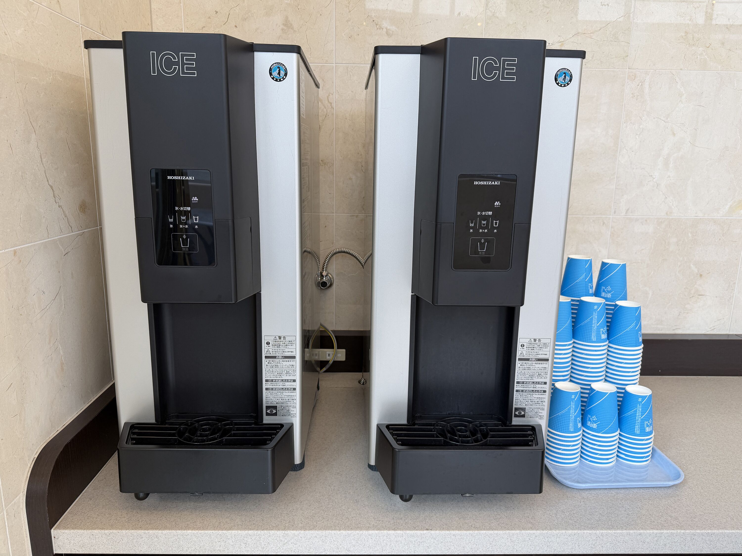 Two ice dispensers labeled ICE, with a stack of blue disposable cups beside them.