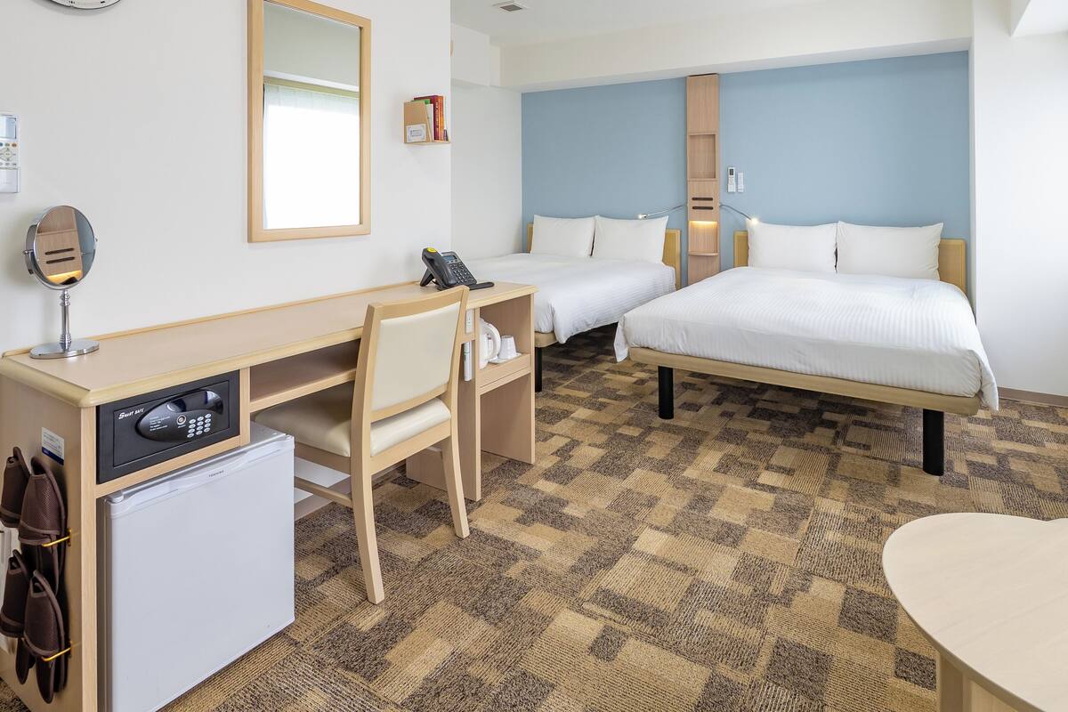 A hotel room with a desk, two beds, a mirror, a clock, and a small refrigerator.
