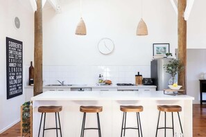 A modern kitchen with a central island, bar stools, hanging pendant lights, and a clock on the wall. - The Deck Sorrento (Sorrento)