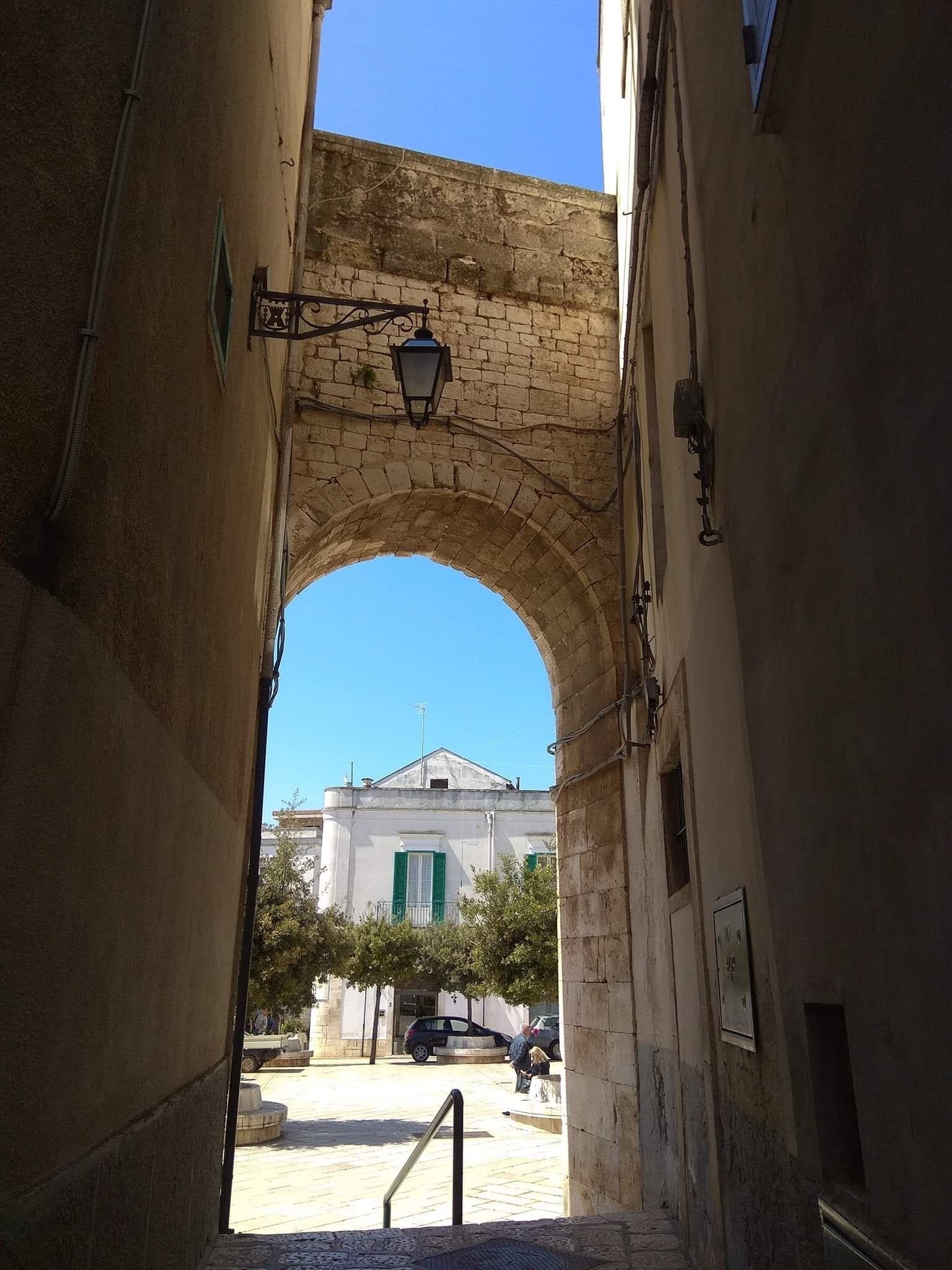 Ein gewölbter Gang mit einer Straßenlaterne und einem Gebäude im Hintergrund, dessen Fassade weiß und die Fensterläden grün sind.