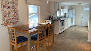 Una cocina con armarios blancos, estufa y pileta. Se observa una mesa de comedor con sillas y un adorno de conchas marinas en la pared.