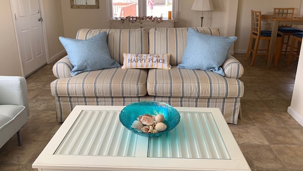 Un sofá a rayas con dos almohadas azules, una mesa de centro de vidrio con un tazón azul y un comedor con sillas de madera y una mesa.