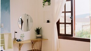 Ein Zimmer mit einem Holztisch, einem Stuhl, einem runden Spiegel, einer Topfpflanze, einem kleinen Regal mit einer Flasche und einem Fenster mit Vorhängen.