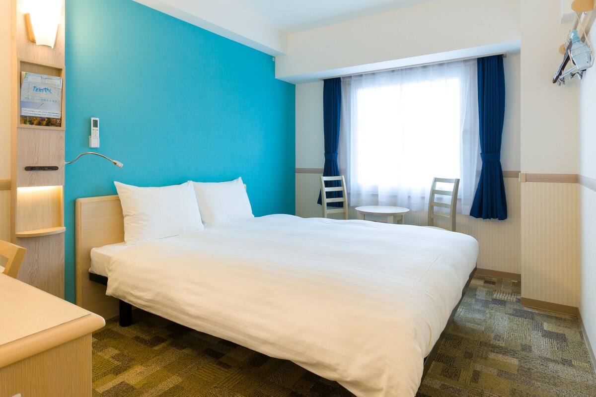 Une chambre d’hôtel avec un grand lit, une table de chevet, un petit bureau, une chaise, une fenêtre avec des rideaux et une lampe fixée au mur.