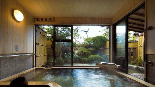 Un onsen japonés tradicional con interior de madera, una puerta de cristal que da a un jardín y un banco de piedra.