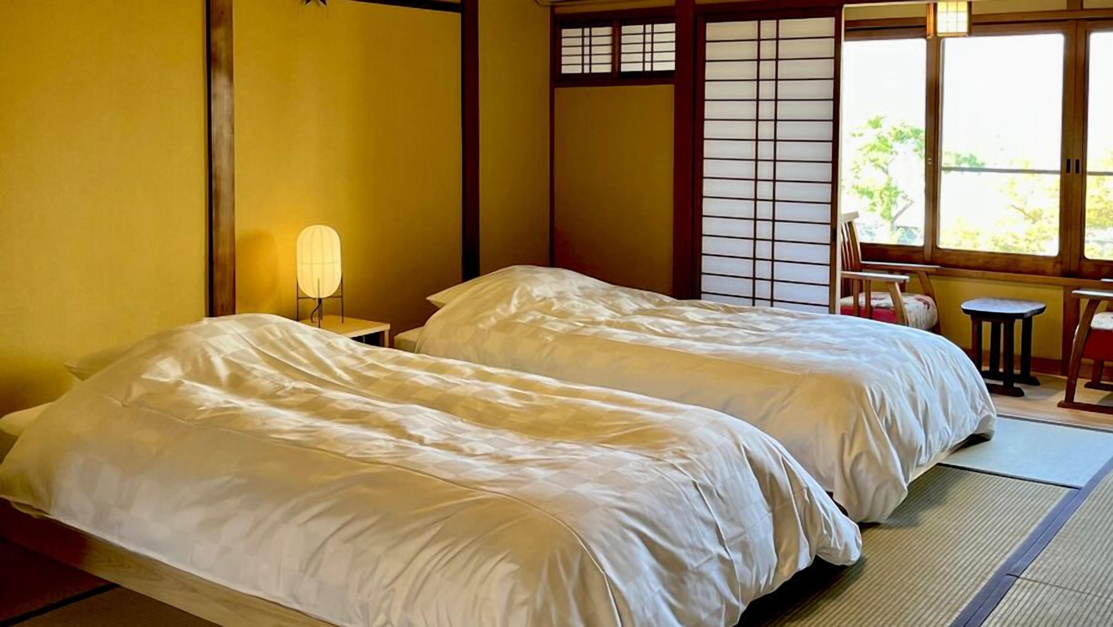 Um quarto japonês tradicional com tatame, duas camas com roupa de cama branca, um biombô shoji e uma pequena mesa com uma cadeira.