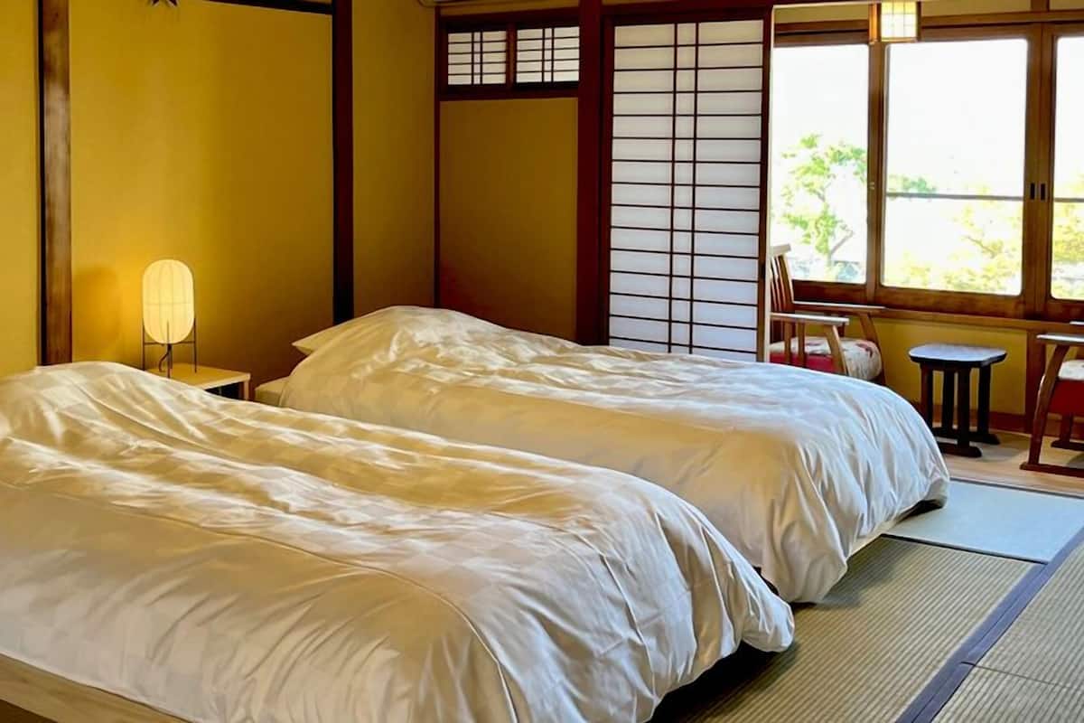Um quarto japonês tradicional com tatame, duas camas com roupa de cama branca, um biombô shoji e uma pequena mesa com uma cadeira.