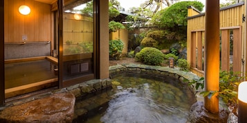 Un onsen japonés tradicional con un balneario de piedra, paredes de madera y un pequeño jardín con rocas y plantas.