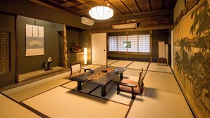 Una habitación japonesa tradicional con suelo de tatami, una mesa baja con cojines, una lámpara colgante y un ventanal con celosías shoji.