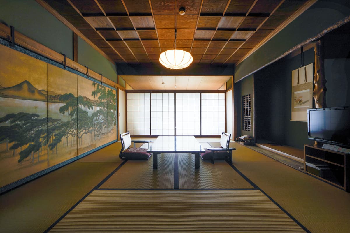 Um quarto japonês tradicional com tatames, uma mesa baixa com almofadas, uma porta deslizante e uma televisão sobre um suporte.