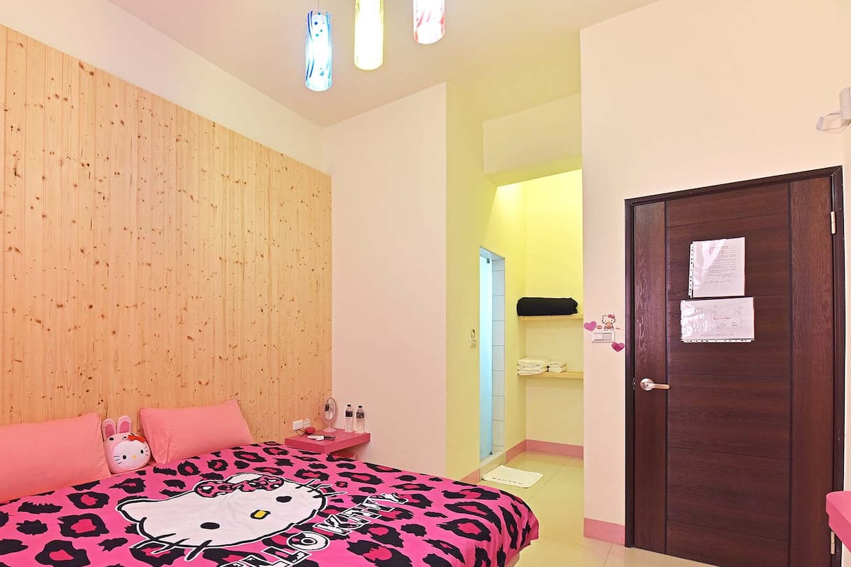 Ein Schlafzimmer mit einem Holz-Kopfteil, einer pinkfarbenen Hello Kitty Bettdecke, einem Nachttisch mit einem Hello Kitty Kissen und einer Tür mit Papieren darauf.