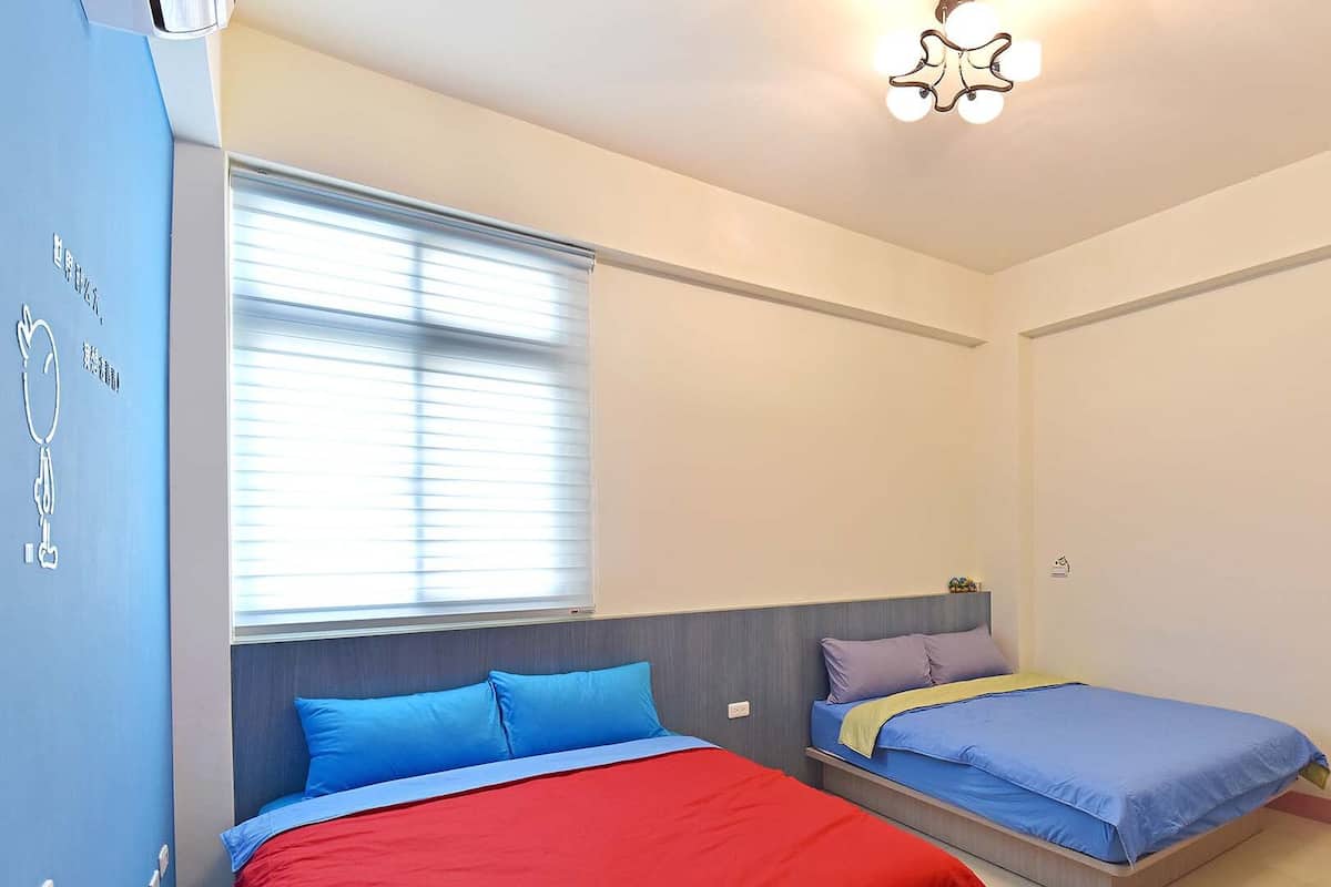 Ein Schlafzimmer mit zwei Betten, einem Fenster mit Jalousien, einer Deckenleuchte und einem kleinen Schreibtisch mit Stuhl.