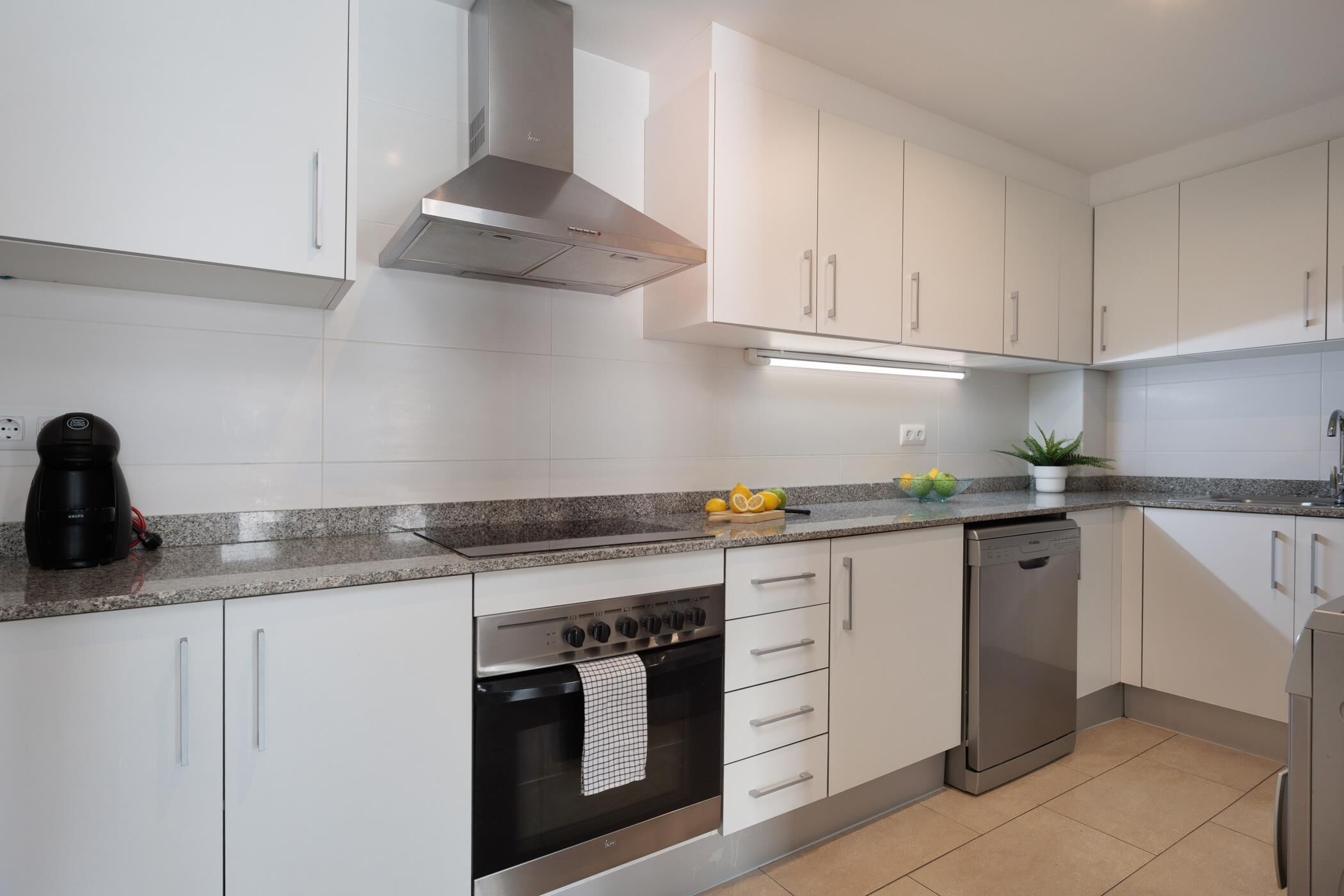 A modern kitchen with white cabinets, a stainless steel range hood, a built-in oven, a dishwasher, and a granite countertop.