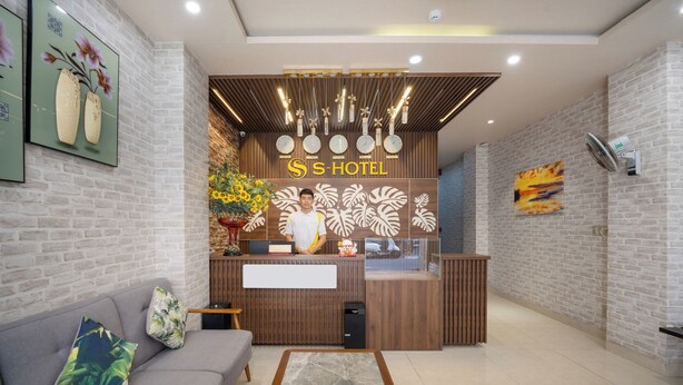 A hotel reception area with a wooden reception desk, a receptionist, and a sofa with decorative pillows. - S HOTEL BEACH DA NANG (Da Nang)