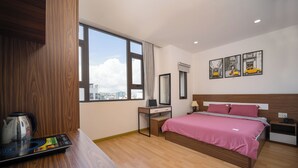A modern hotel room with a large window, a bed with pink bedding, a desk with a laptop, a coffee maker, and artwork on the wall. - S HOTEL BEACH DA NANG (Da Nang)