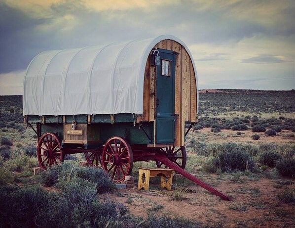 A covered wagon with a wooden structure and red wheels, situated in a desert landscape.