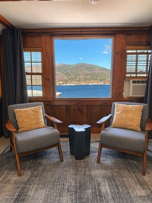 A room with two armchairs, a small black table, and a large window with a view of a lake and hills. - Lake George Halls House  (Lake George)
