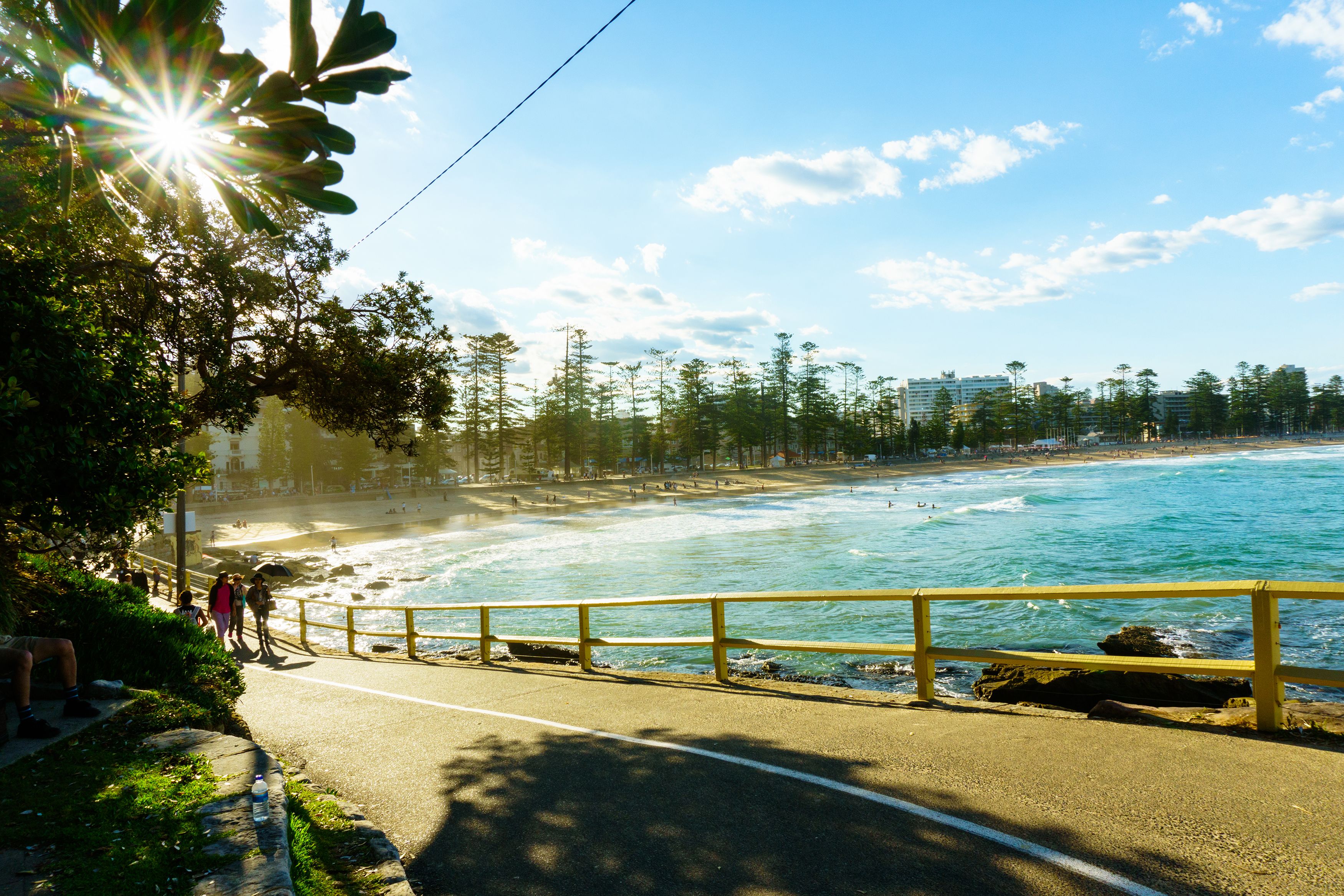 A coastal road with a yellow railing, leading to a beach with waves and a clear sky.