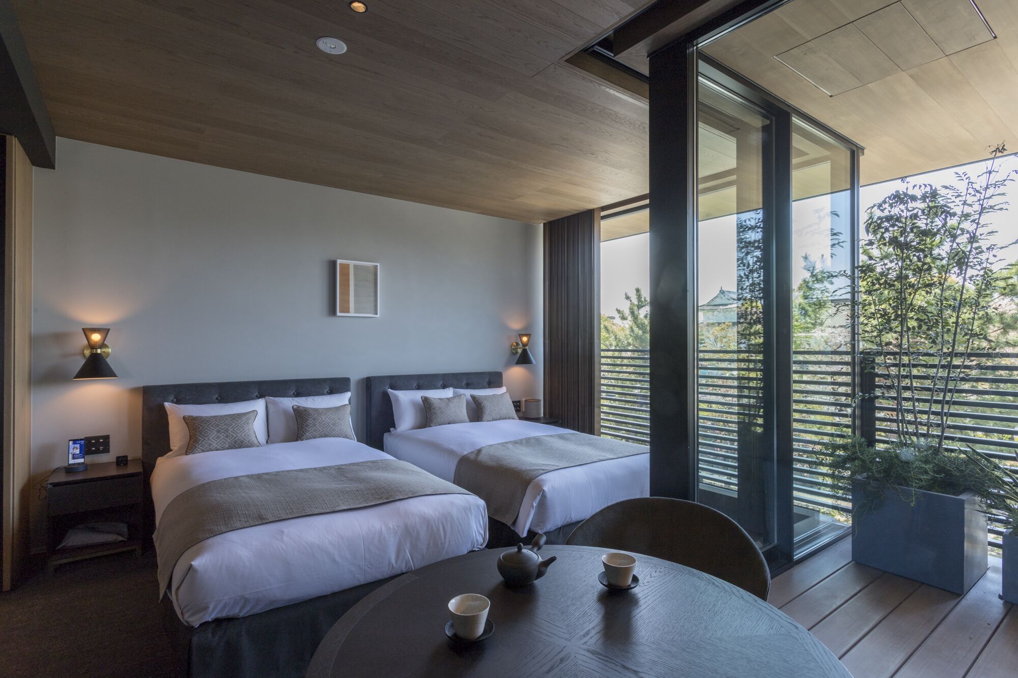 A modern hotel room with two beds, a small table with cups and a teapot, and a balcony with a view of trees.