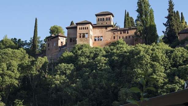 Un edificio histórico con múltiples torres y un muro fortificado, rodeado de abundante vegetación.
