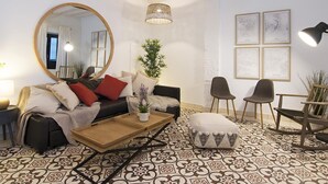 Una sala de estar con sofá, sillas, una mesa de centro, un espejo redondo, una lámpara colgante y obras de arte enmarcadas en la pared.