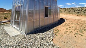 A transparent greenhouse structure with a metal roof, situated in a desert environment with gravel ground and sparse vegetation.